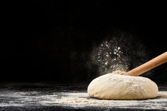 Rustic bread dough being kneaded on a dark surface with flour dust in the air