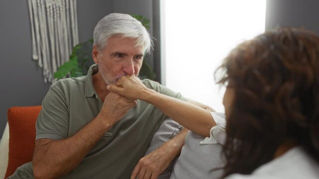 Man kissing woman's hand in intimate gesture, sitting together in cozy living room with warm lighting, depicting love and connection in an indoor setting.