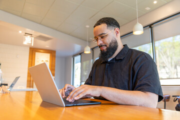 An African American man focuses on his laptop at a wooden table in a bright coffee shop, where natural light streams through large windows, creating a cozy atmosphere for creativity