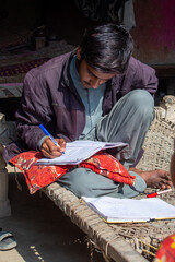 Fototapeta premium Pakistani Village Boy Studying on a Charpai | Rural Education Concept | Focused Young Student Writing in Notebook Outdoors