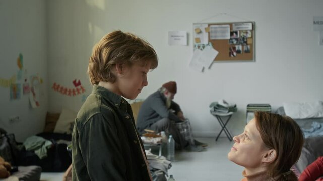 Medium side shot of young Caucasian mom comforting son traumatized by war or disaster in refugee shelter, stroking hair, buttoning shirt, talking, encouraging, then both hugging