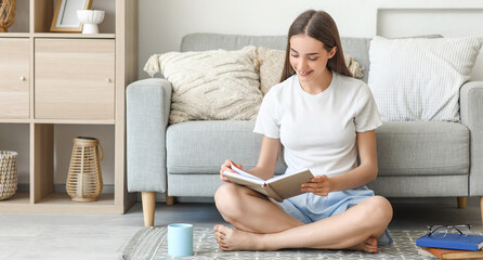 Beautiful young happy woman with cup of tea reading book on floor at home