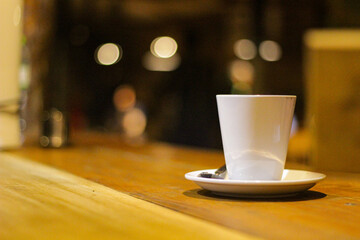 A white coffee cup sits on a wooden table with a saucer underneath it. The cup is filled with coffee and a spoon is resting on the saucer. The scene is simple and uncluttered.