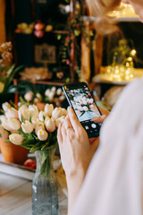 Close-up of person photographing bouquet of white tulips using smartphone in floral arrangement shop. Background features beautiful lighting and floral decor, creating warm and inviting atmosphere.