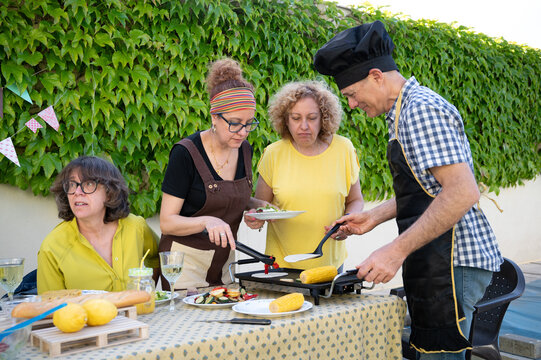 Group of four adults preparing a homemade meal outdoors, grilling tortillas and corn on a griddle, sharing fresh food and drinks on a decorated table in a backyard setting with greenery.