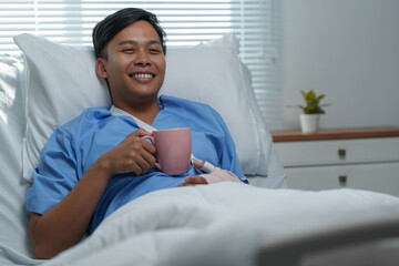 Young asian patient recovering in hospital bed, enjoying a hot beverage