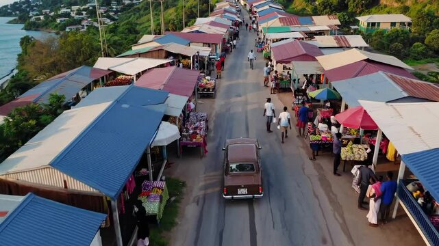 Aerial video shot of a coastal village market, showcasing vibrant stalls and a car on a road, with a scenic ocean view on the left.