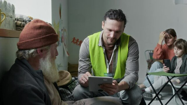 Medium full shot of cheerful Caucasian male social worker in yellow vest sitting on chair next to elderly displaced man with beard, asking questions, filling form, while responding to natural disaster