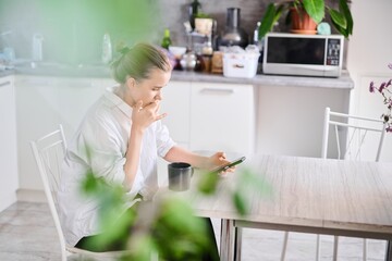 Cute teenage schoolgirl drinks coffee for breakfast and checks email, looks at smartphone screen