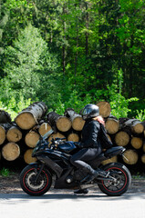 Biker girl in a black jacket on a motorcycle looking at the straight. Motorcyclist in outfit with motorcycle in sunglasses and jeans in summer. Side view. with tree logs in the background