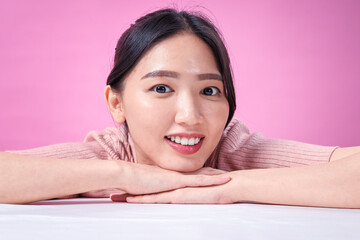Obraz premium A young Asian woman with dark hair in a bun, wearing a ribbed pink short-sleeve top, rests her head on her crossed arms while smiling warmly against a solid pink background, creating a relaxed
