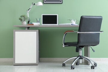Stylish workplace with armchair, modern laptop, lamp and houseplant near green wall in office