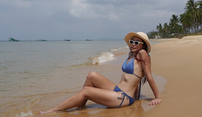 Asian woman  lounging on a sandy beach, wearing a blue bikini set and a straw hat with white...