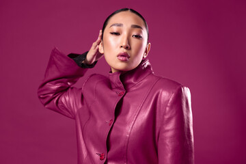 Fashion asian brunette woman in purple leather jacket posing with hands on head against purple background