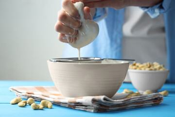Woman straining cashew milk into bowl at light blue wooden table with nuts indoors, closeup