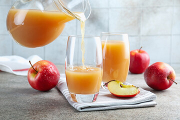 Pouring refreshing apple juice from jug into glass at grey textured table, closeup