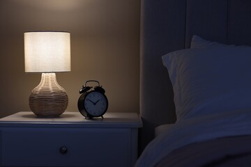 Glowing lamp and alarm clock on nightstand in bedroom