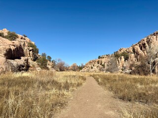 Dirt trail through dry grassland, surrounded by rocky cliffs, blue sky