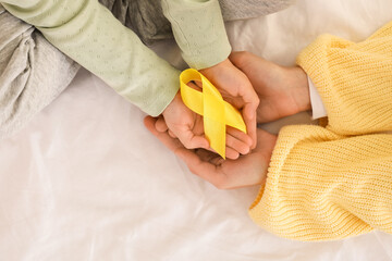 Little girl after chemotherapy with yellow ribbon and mother on bed, closeup. Childhood cancer awareness concept