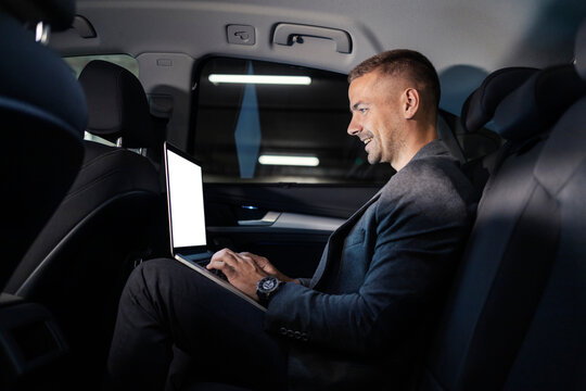 Young busy executive is working on his laptop in a car at backseat and smiling. - Powered by Adobe