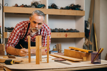 Senior carpenter in plaid shirt and overalls inspects a wooden stool in a workshop, surrounded by tools and materials. Workshop wall with tools visible in the background, showcasing craftsmanship