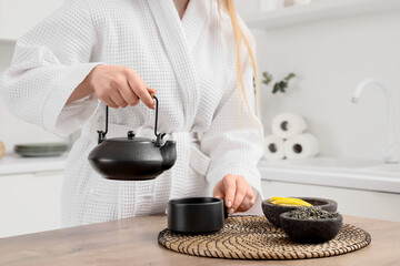 Young woman in bathrobe with teapot, cup and dry tea leaves in kitchen, closeup