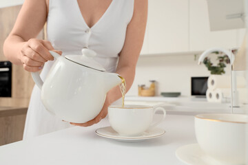 Young woman pouring tasty tea from teapot into cup on table in kitchen, closeup