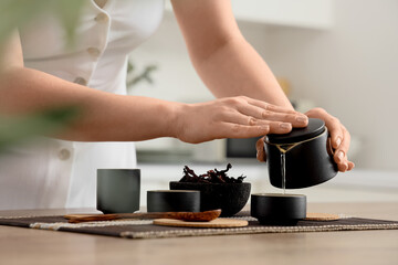 Young woman pouring tasty tea from teapot into cup on table in kitchen, closeup