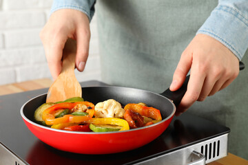 Woman frying vegetables in pan on stove at table indoors, closeup