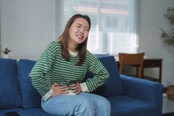 Obraz premium Young woman suffering from abdominal pain while sitting on sofa at home