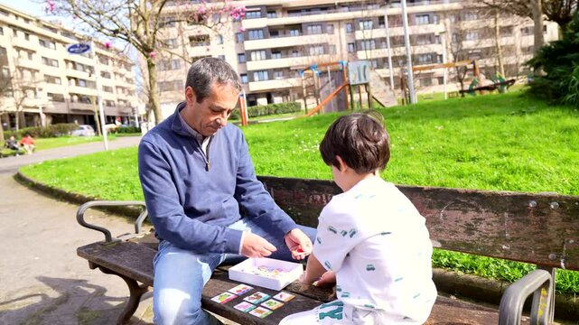 Elderly grandfather teaching card game to young grandson while sitting together on sunny park bench, sharing heartwarming moment of intergenerational bonding and family connection