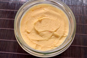 A glass jar displays creamy hummus with a smooth surface and vibrant yellow hue. It sits on a textured surface, showcasing its rich consistency, perfect for dipping or spreading
