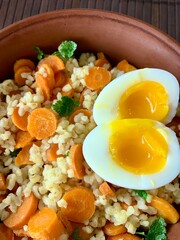 A vibrant dish featuring cooked barley tossed with sliced carrots, topped with two soft-boiled eggs and fresh herbs. Perfect for a healthy meal