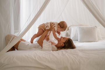 The young woman mother lies on the white bed and lifts her little daughter on the hands. They play and laugh. Maternity leave lifestyle. The woman tenderly kisses her infant daughter. Mothers day.