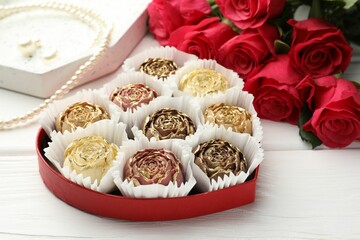 Flower shaped chocolate bonbons in box, red roses and pearl jewelry on white wooden table, closeup
