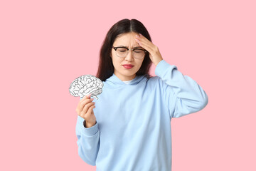 Young Asian woman with paper brain suffering from headache on pink background. Brain Injury Awareness Month