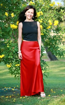 Beautiful brunette woman 51 years old in a satin red long skirt posing against the background of a flowering tree in Goa India