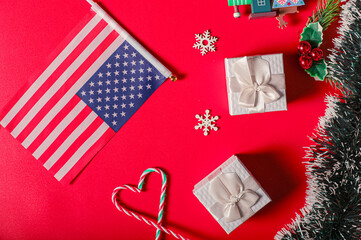 Fototapeta premium A Boxing Day preparation flat lay featuring a U.S. flag, white gift boxes, candy canes shaped like a heart, snowflake decorations, holly, and Christmas greenery on a vibrant red background.