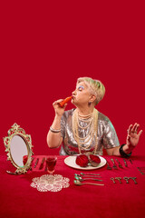 Elderly woman using carrot as lipstick, sitting at red table with surreal meal of beets, wearing pearls and silver top. Vibrant lifestyle. Pop art photography. Concept of humor and elegance, self care