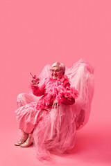 Stylish elderly woman in extravagant pink outfit sitting on pink chair, holding glass with pink ice cream. Bold self-expression. Pop art photography. Concept of eccentric lifestyle and quirky fashion