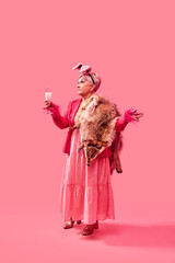 Glamorous elderly woman in pink outfit standing against pink background, raising glass of milk in...