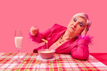 Elderly woman in vibrant pink outfit elegantly holding spoon with cereal, resting head on hand, gazing with calm expression against pink background. Food pop art photography. Concept of fashion