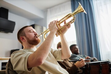 Side view portrait of young man playing golden trumpet while enjoying sports game on TV at home...