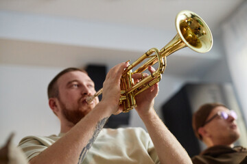 Obraz premium Close up of red haired man playing trumpet enthusiastically while sitting on couch at home with friends, copy space