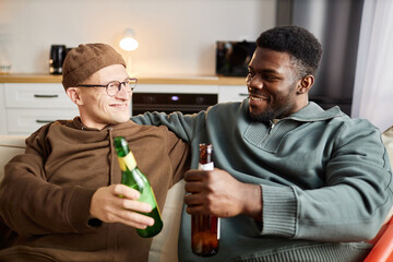 Front view portrait of two smiling male friends clinking beer bottles in cheers while sitting on couch at home and watching TV