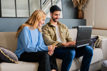 Couple joyfully shopping online together while sitting comfortably on a cozy sofa, sharing laughter and excitement during their engaging digital purchasing experience with a laptop and credit card