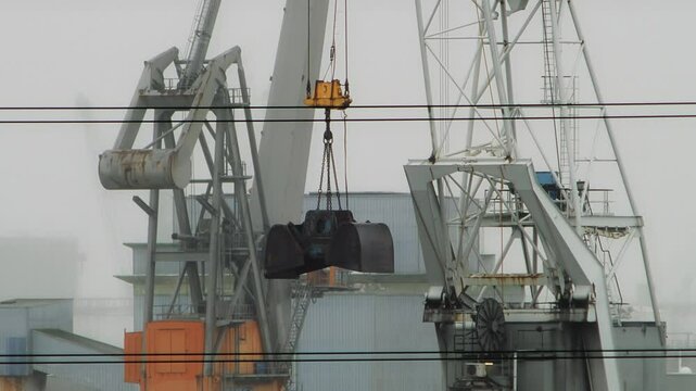 Close-up of a fixed heavy-duty port crane with a clamshell grab bucket swaying in the wind in a foggy harbor