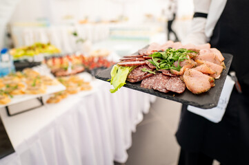 A close-up of a waiter holding a black platter filled with assorted meats and greens, served at an elegantly arranged catering event