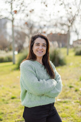 Obraz premium A woman in a soft green sweater and black jeans stands with arms crossed in an urban park, smiling. Warm sunlight enhances the relaxed and confident mood. Nature and self-care concept. Vertical.