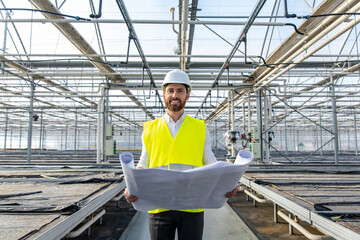 Young bearded engineer in hardhat working with sketches in a greenhouse structure